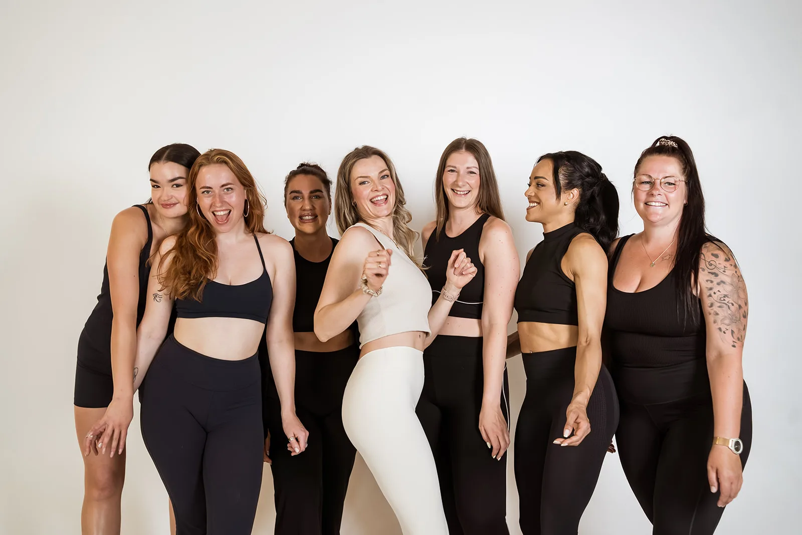 Sieben Frauen in schwarzer und cremefarbener Sportkleidung lachen Arm in Arm vor einer hellen Wand.